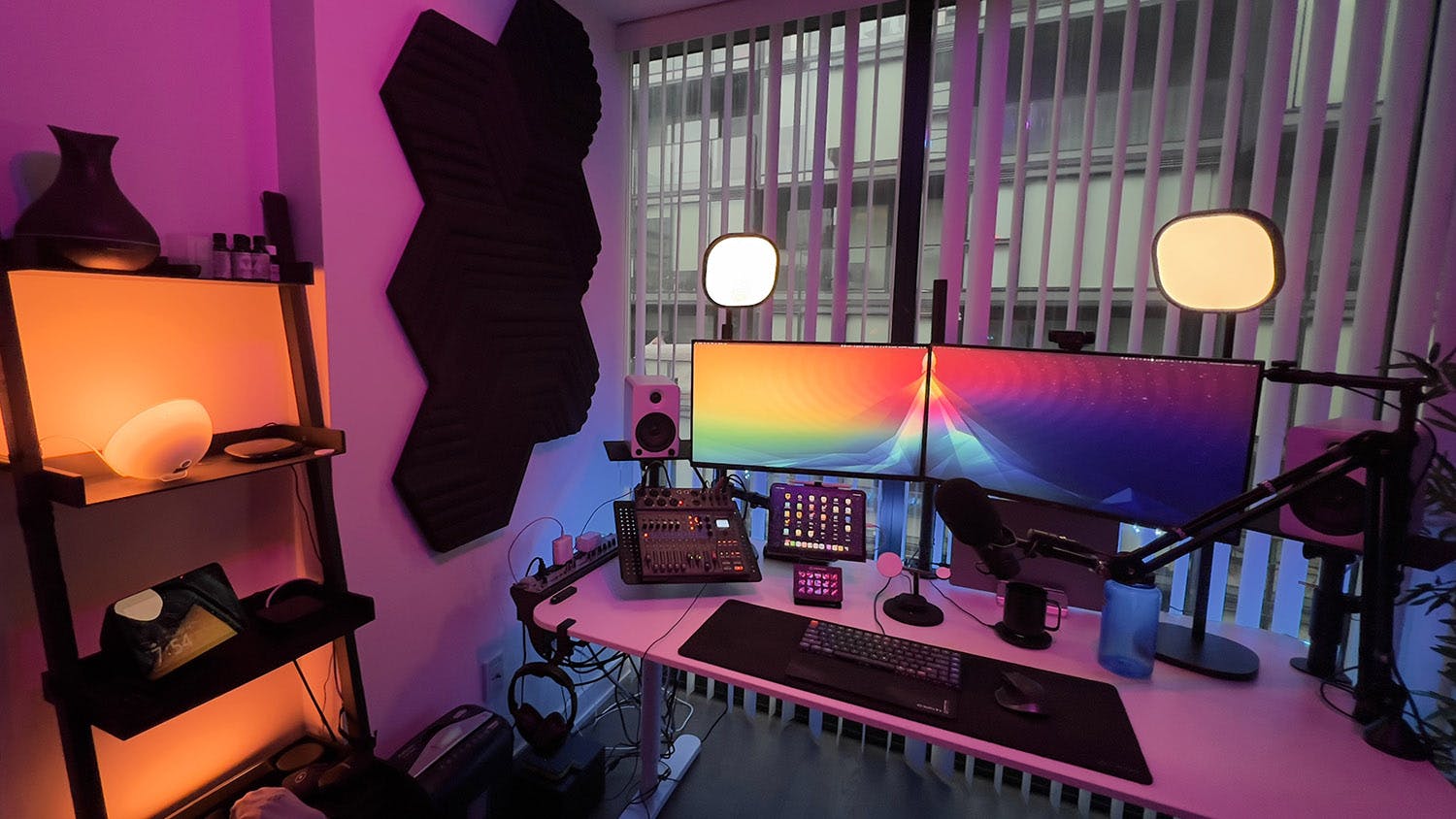 Photo of my desk setup with pink and warm yellow Philips Hue lights. My white desk is composed with 2 screens with a colorful screen background. Two white speakers in each side of the desk. A gray keyboard and a black mouse on a black pad. A microphone on a black arm attached to the right side of the desk.